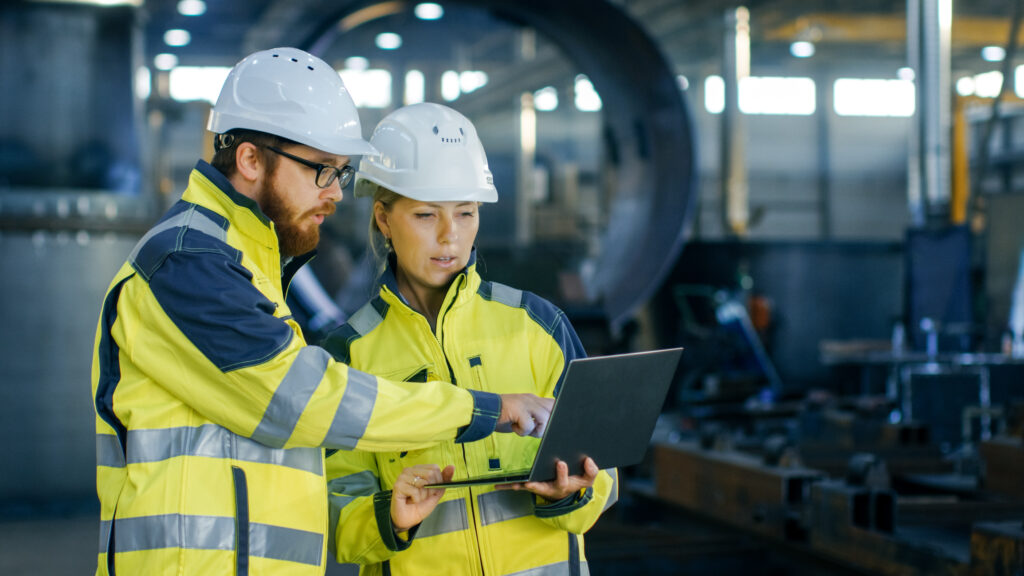 Zwei Mitarbeiter in der Industrie mit gelben Warnwesten besprechen das Bauprojekt am Laptop