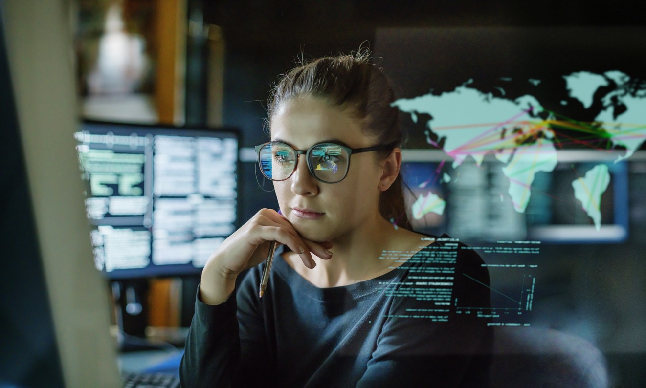 Stockbild einer jungen Frau mit Brille, die in einem dunklen Büro von Computermonitoren umgeben ist. Vor ihr befindet sich ein durchsichtiges Display, das eine Weltkarte mit einigen Daten zeigt.